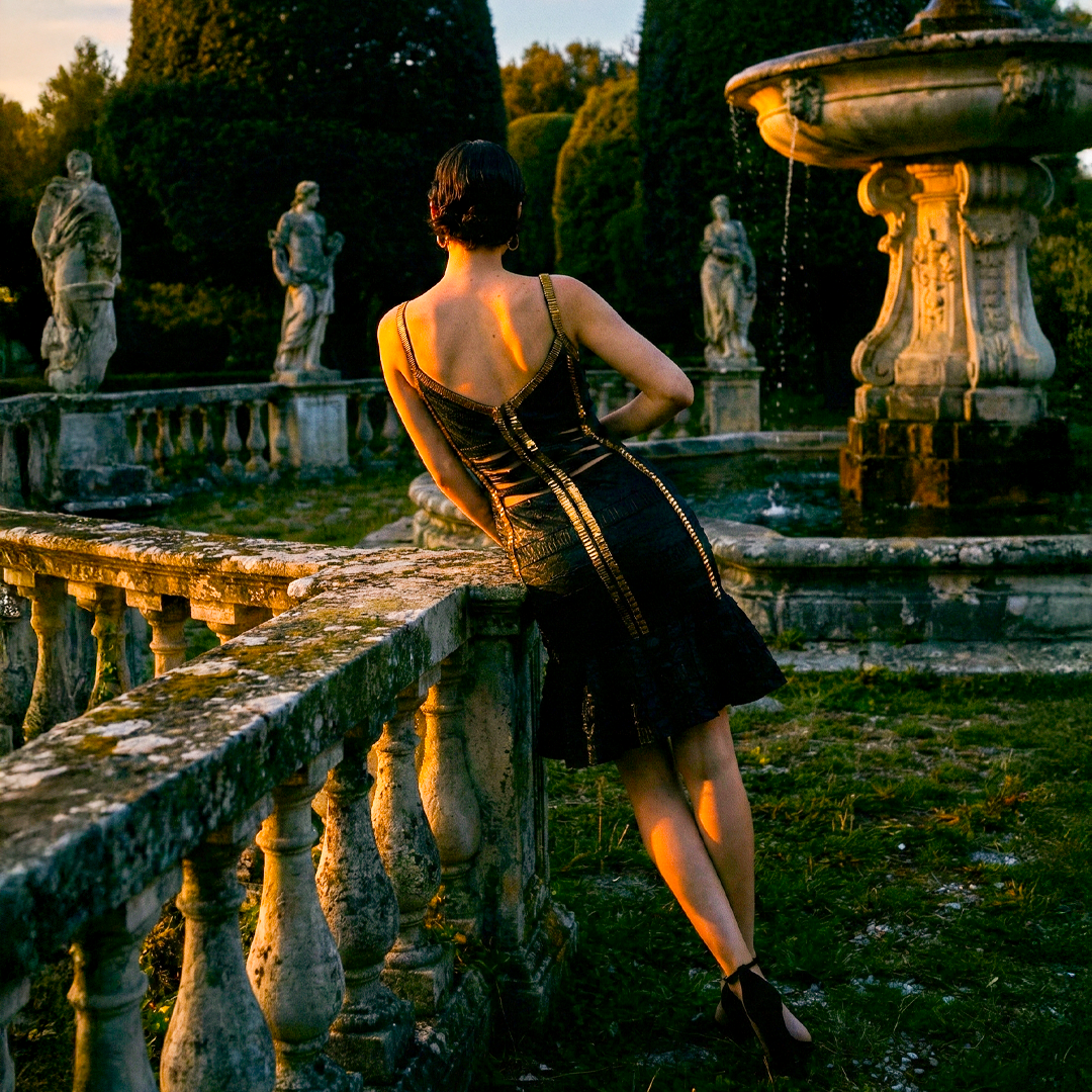 Image of Back view of woman wearing black bandage dress with gold trim and tiered ruffle mermaid hem.