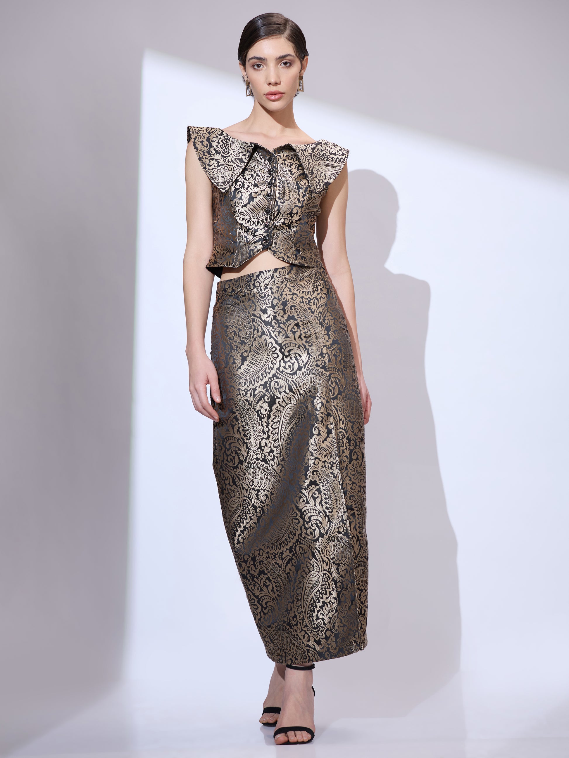 Standing straight in a black and gold metallic brocade co-ord set with a cropped top and a floor-length pencil skirt