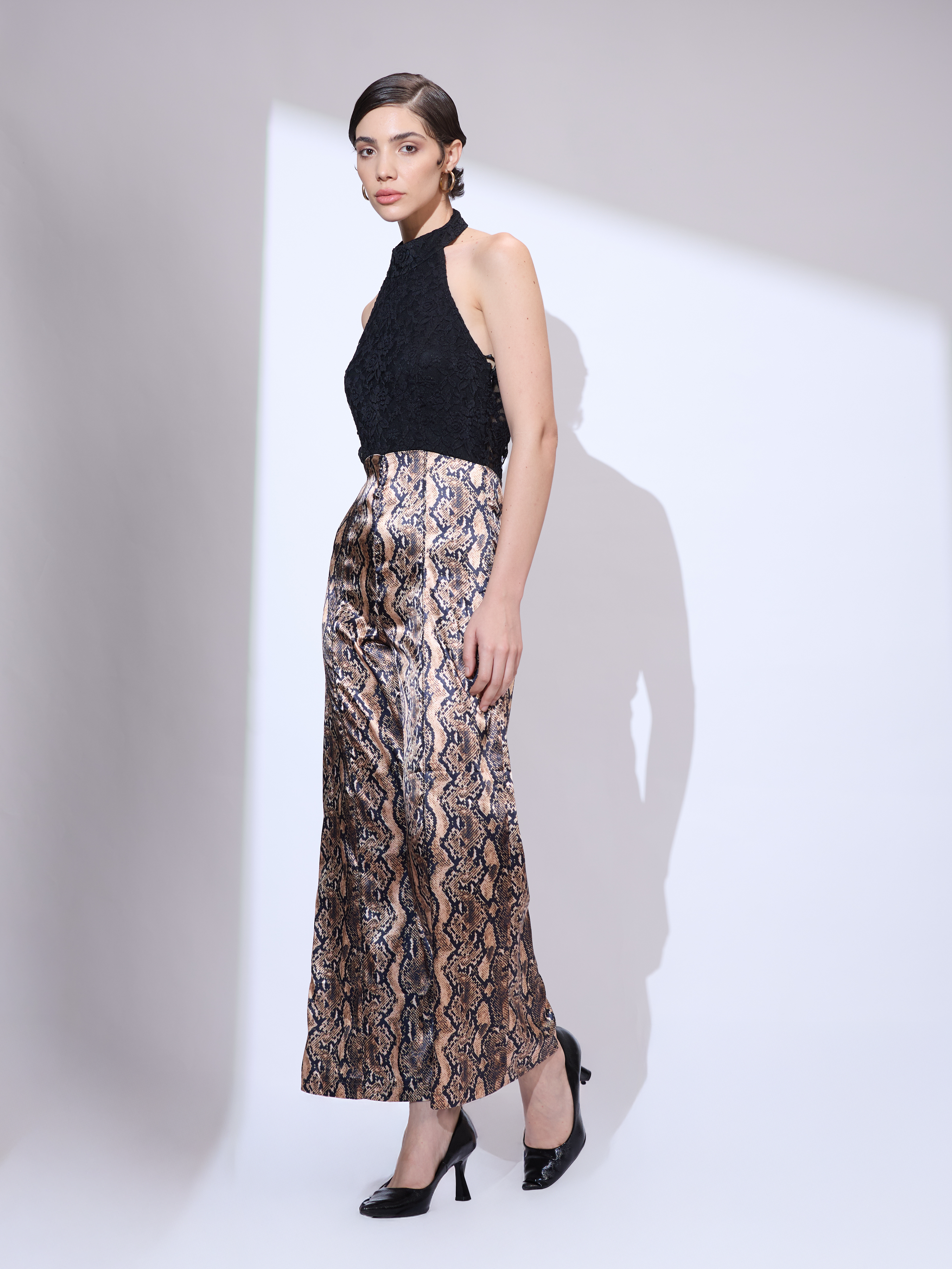 Image of Standing slightly to the side and looking forward in a black and gold metallic snakeskin halter neck jumpsuit with a wide-leg silhouette.