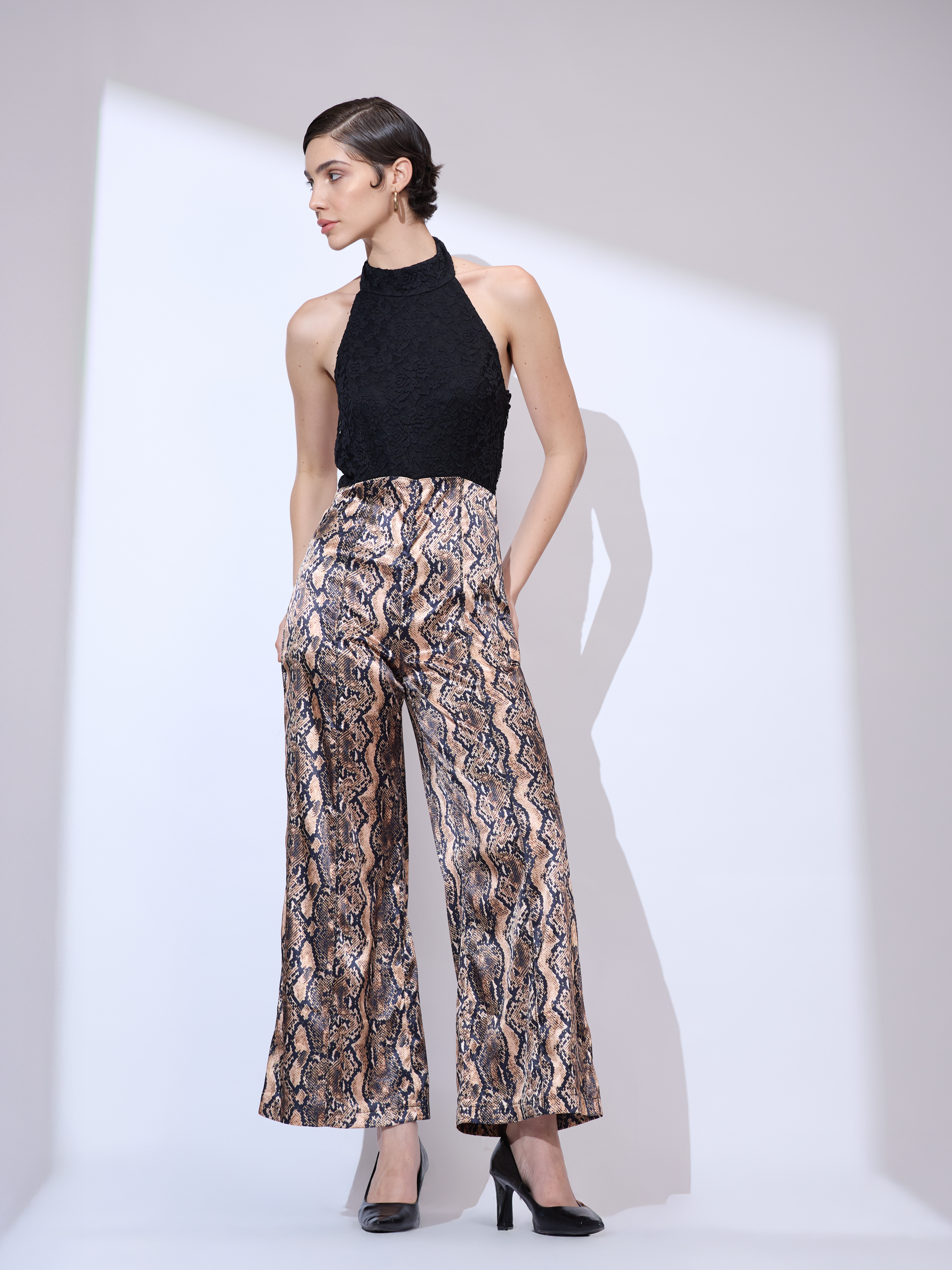 Image of Standing straight and looking left with hands in pockets in a black and gold metallic snakeskin wide-leg jumpsuit with a halter neckline.