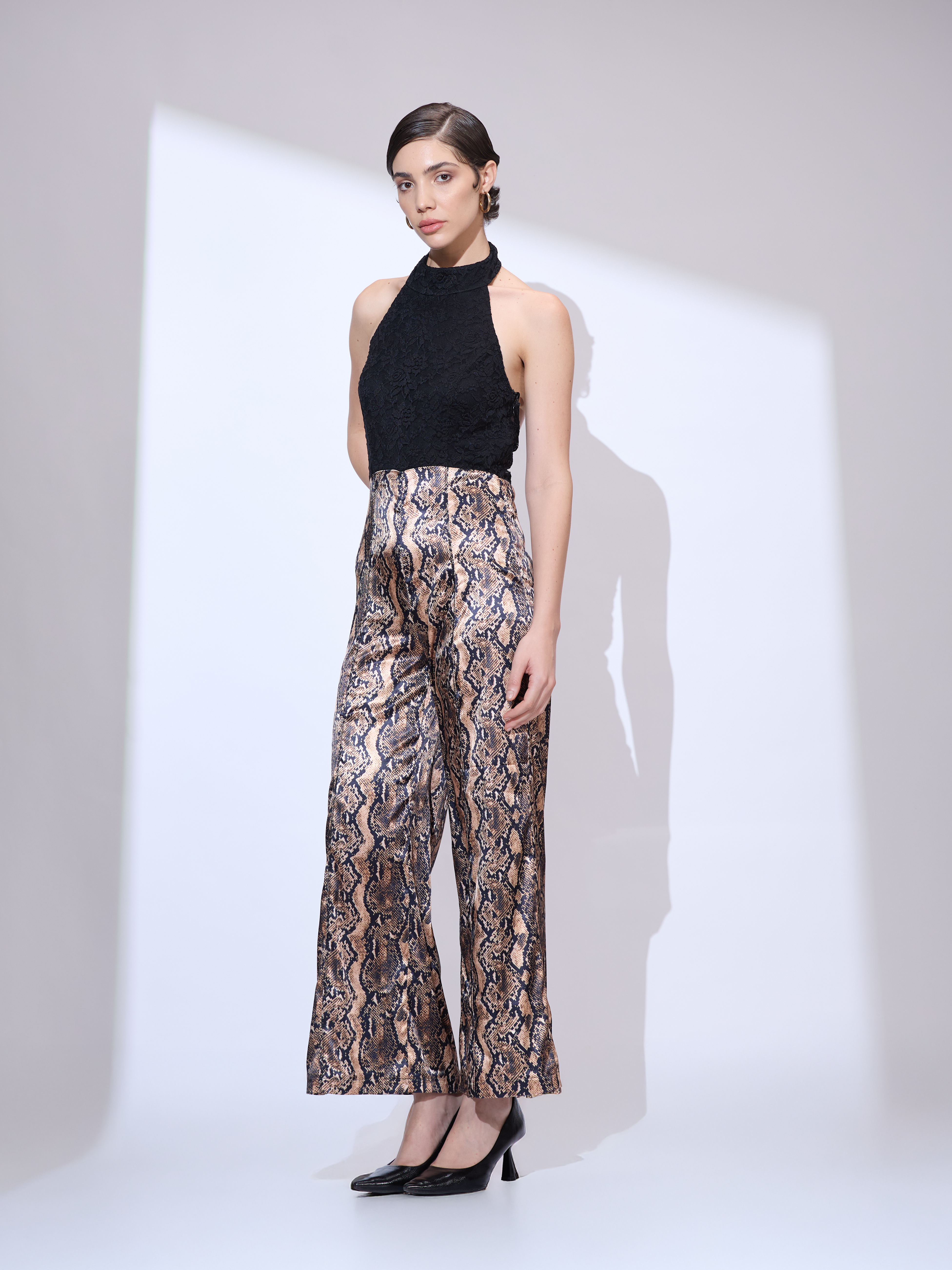 Image of Standing slightly to the side and facing forward in a black and gold metallic snakeskin wide-leg jumpsuit with a halter neckline.