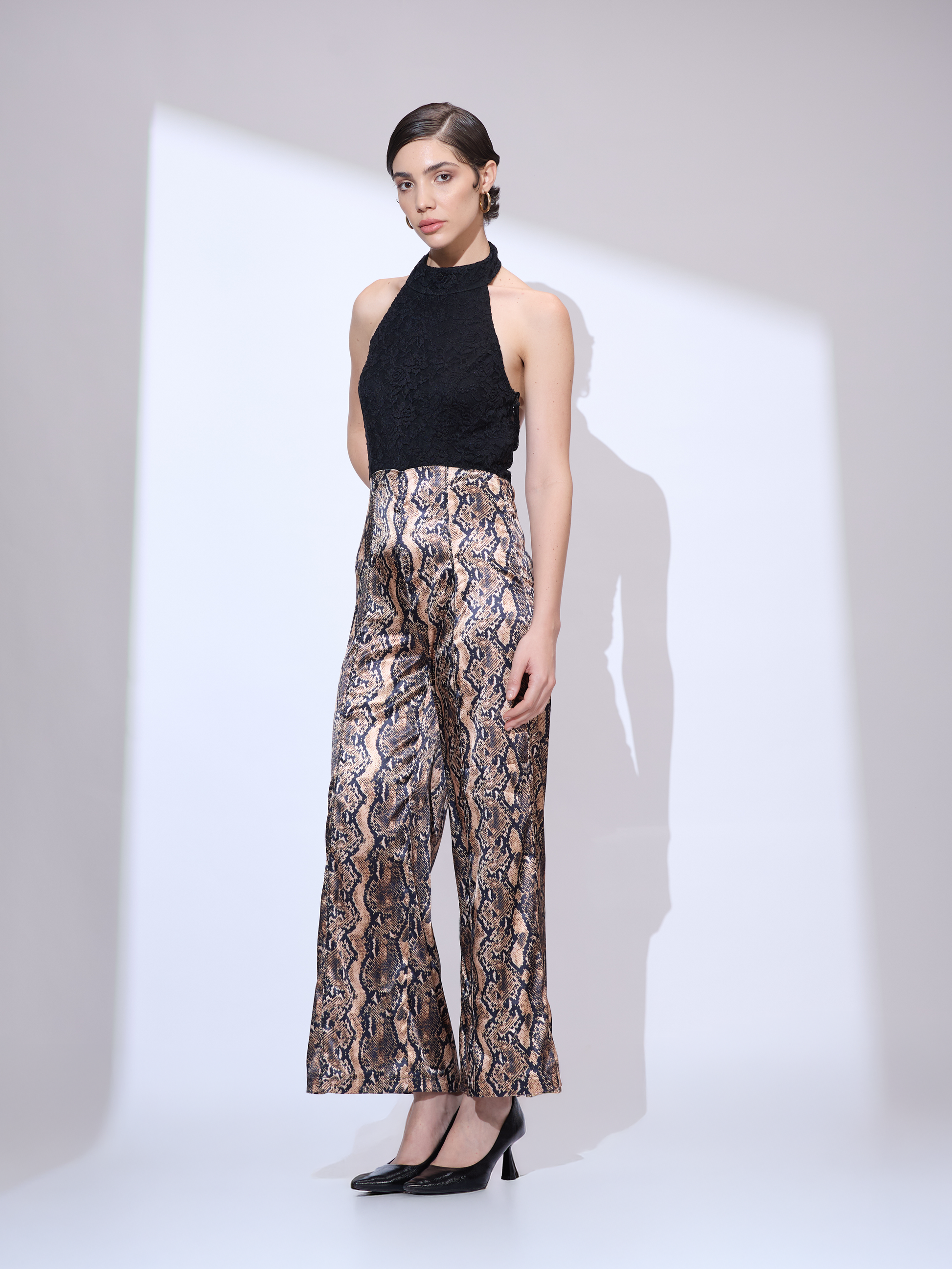 Standing slightly to the side and facing forward in a black and gold metallic snakeskin wide-leg jumpsuit with a halter neckline.