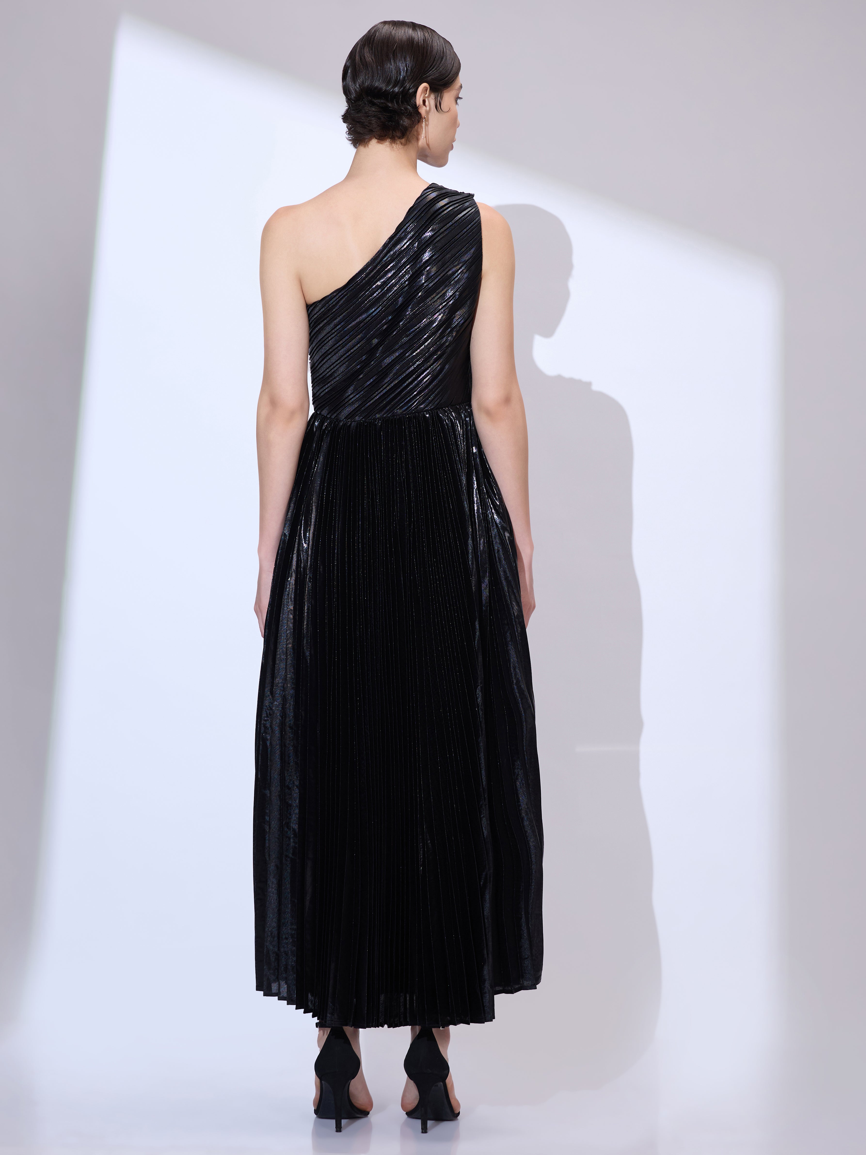Image of Back view of woman wearing a metallic pleated black one shoulder dress with flowing scarf drape