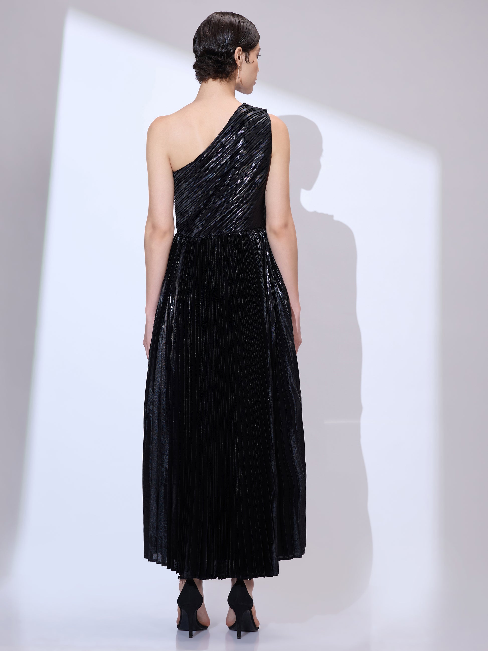 Back view of woman wearing a metallic pleated black one shoulder dress with flowing scarf drape