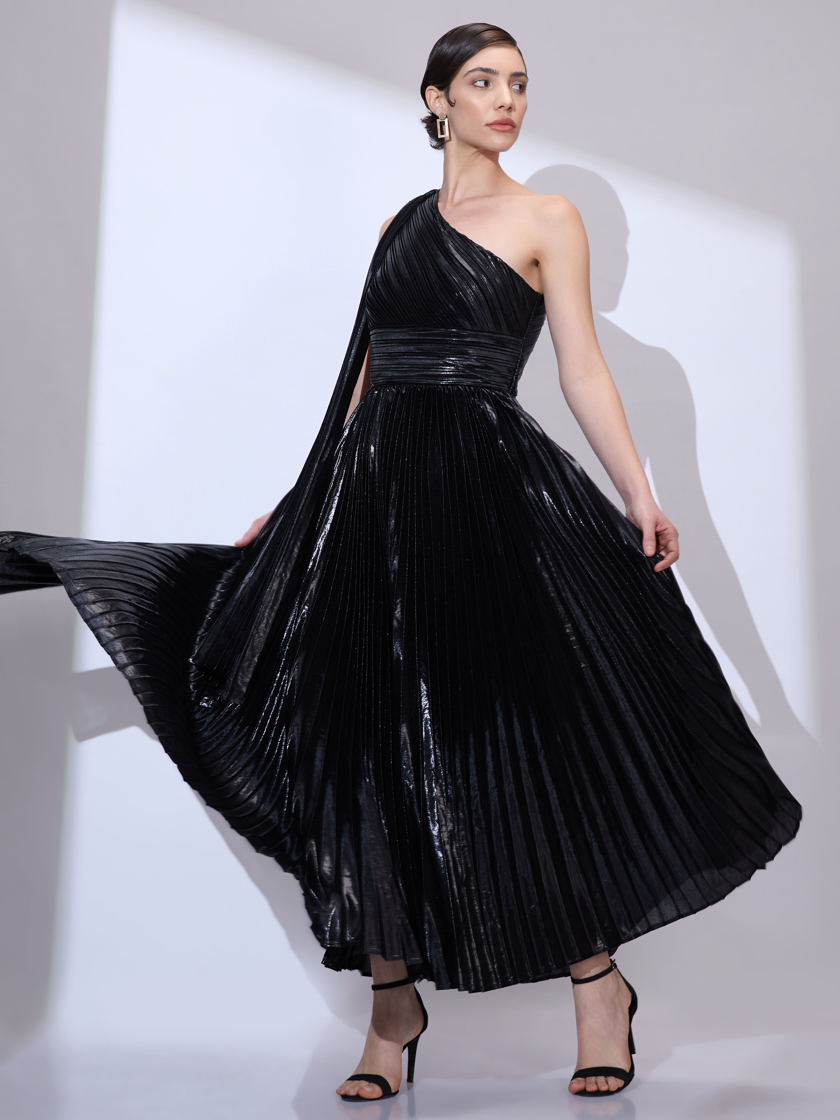 Image of Holding the dress with both hands wearing a metallic pleated black one-shoulder dress with a flowing scarf drape.