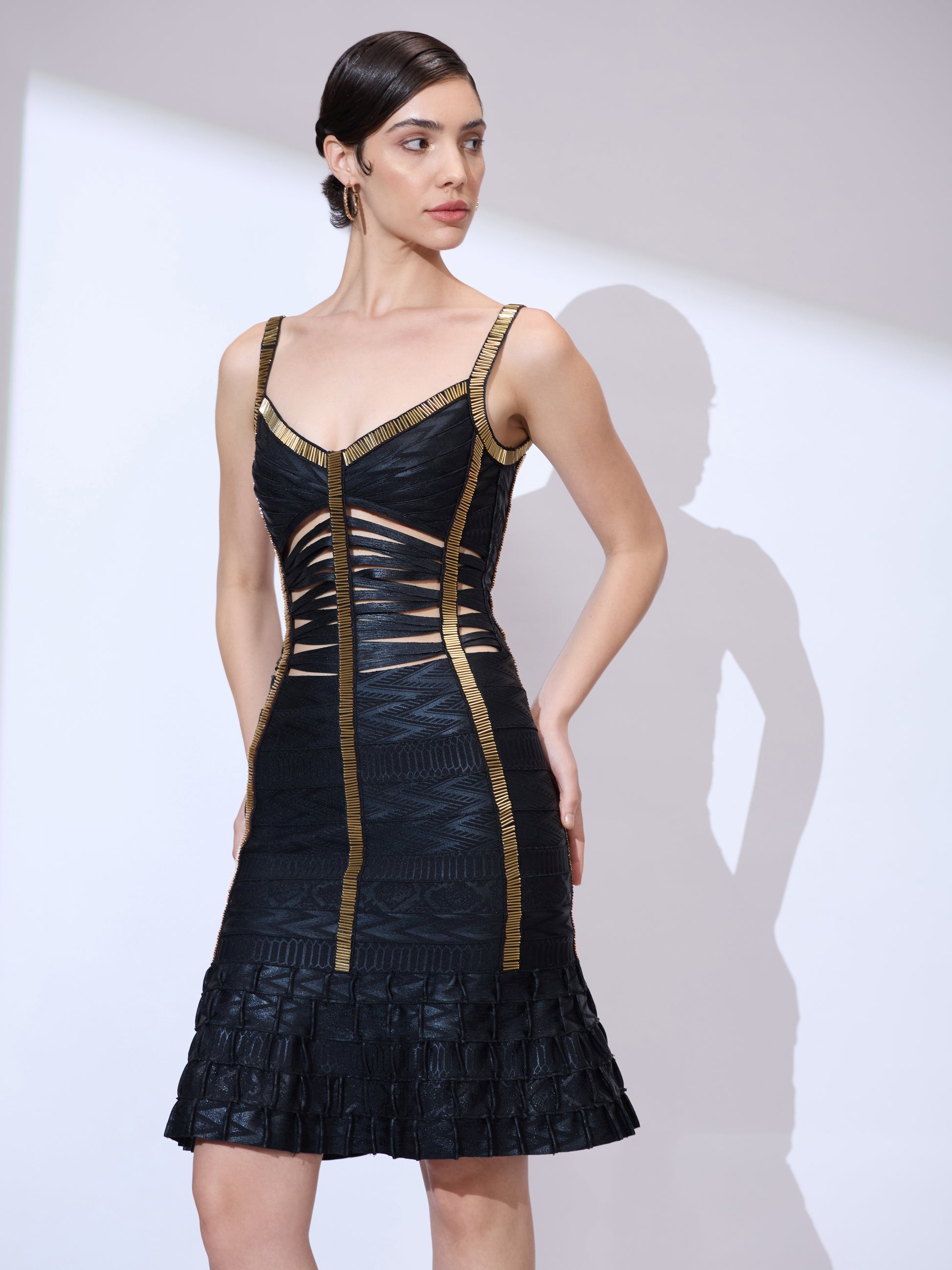 Looking right with hands on waist wearing a ruffle hem dress with gold trim and bodycon silhouette.