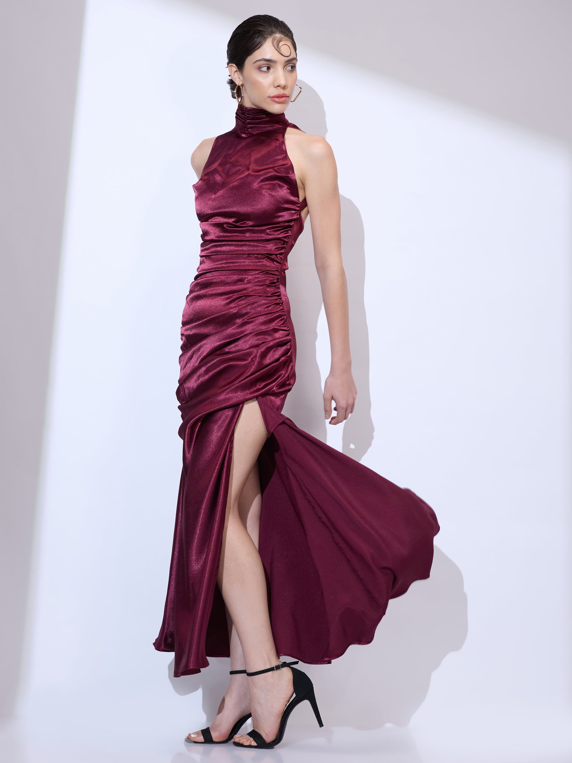 Walking in a burgundy silk satin halter maxi dress with an elegant cowl neckline, ruched detailing, and flowing floor-length silhouette.