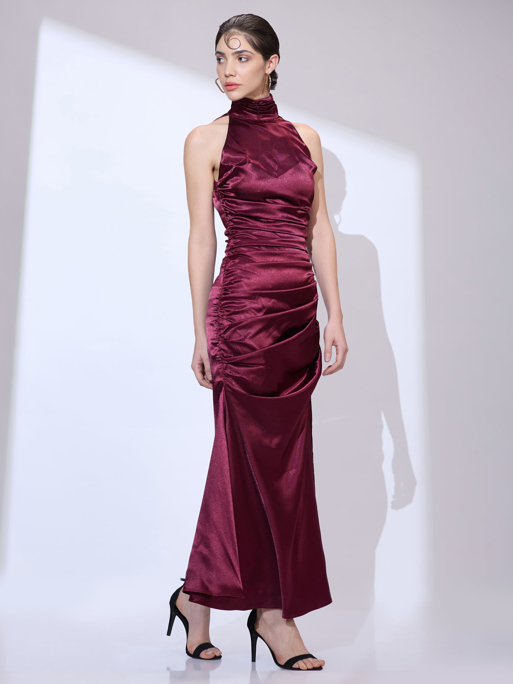 Standing straight and looking left in a burgundy silk satin halter maxi dress with an elegant cowl neckline and ruched hip detail.