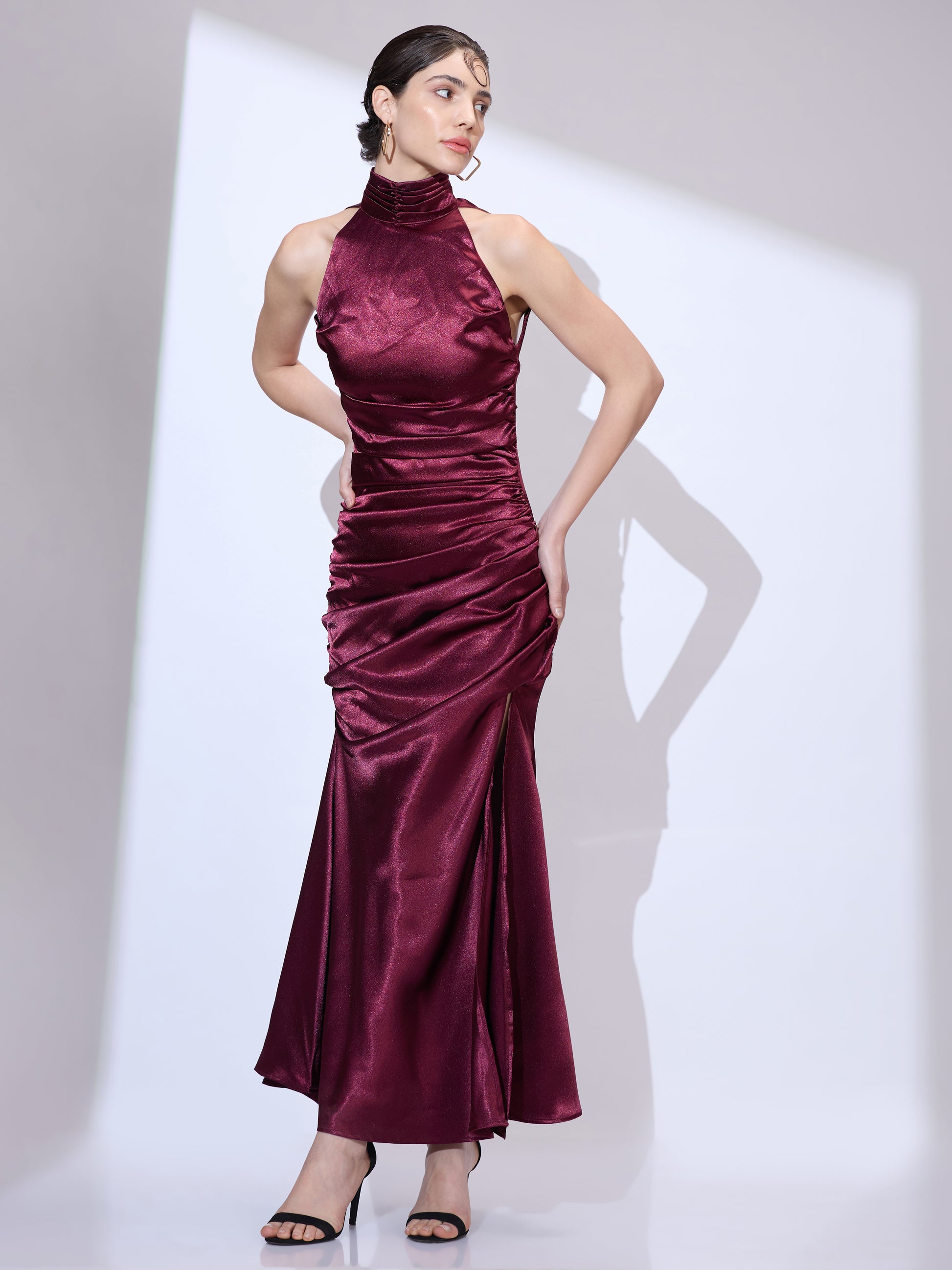 Standing in a burgundy silk satin halter neck maxi dress with an elegant cowl neckline and ruched hip detailing.