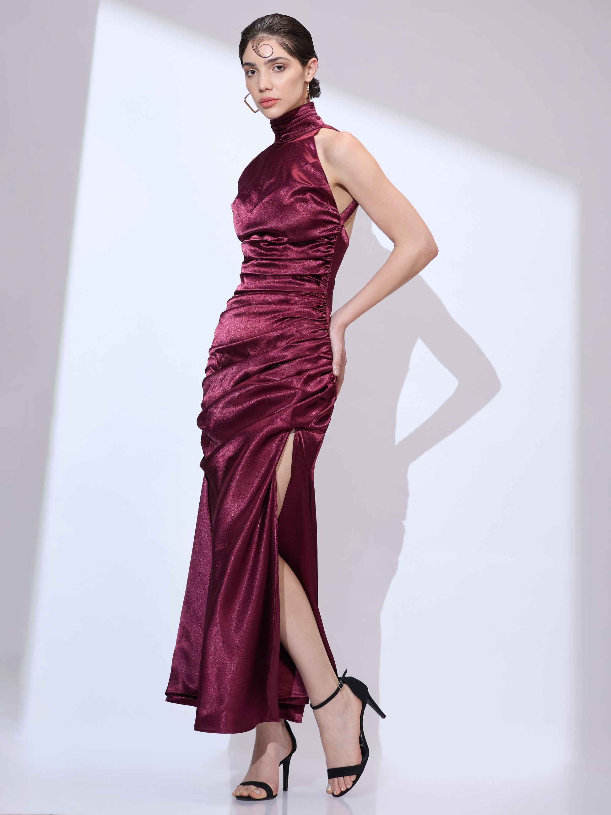 Standing slightly to the left with hands on waist in a burgundy silk satin halter neck maxi dress with an elegant cowl neckline and ruched detailing.