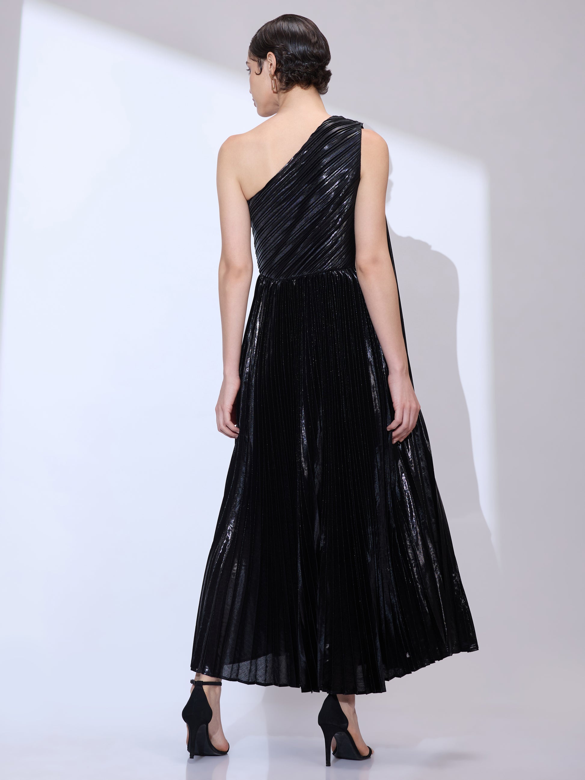 Back view of woman wearing an asymmetrical neckline gown in metallic pleated fabric with a flowing shoulder scarf