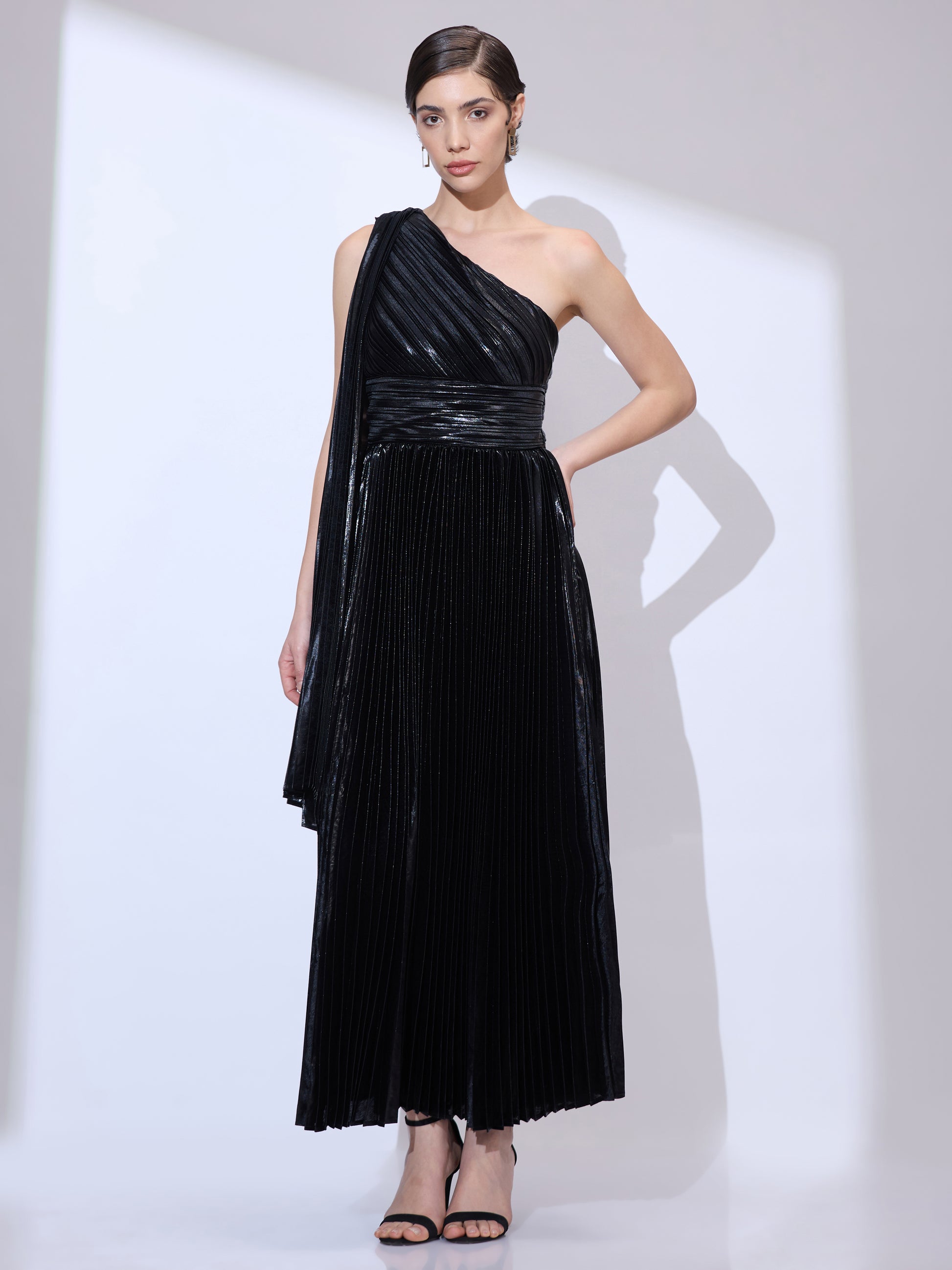 Standing straight with hands on waist wearing a metallic pleated one-shoulder dress with a flowing scarf drape.