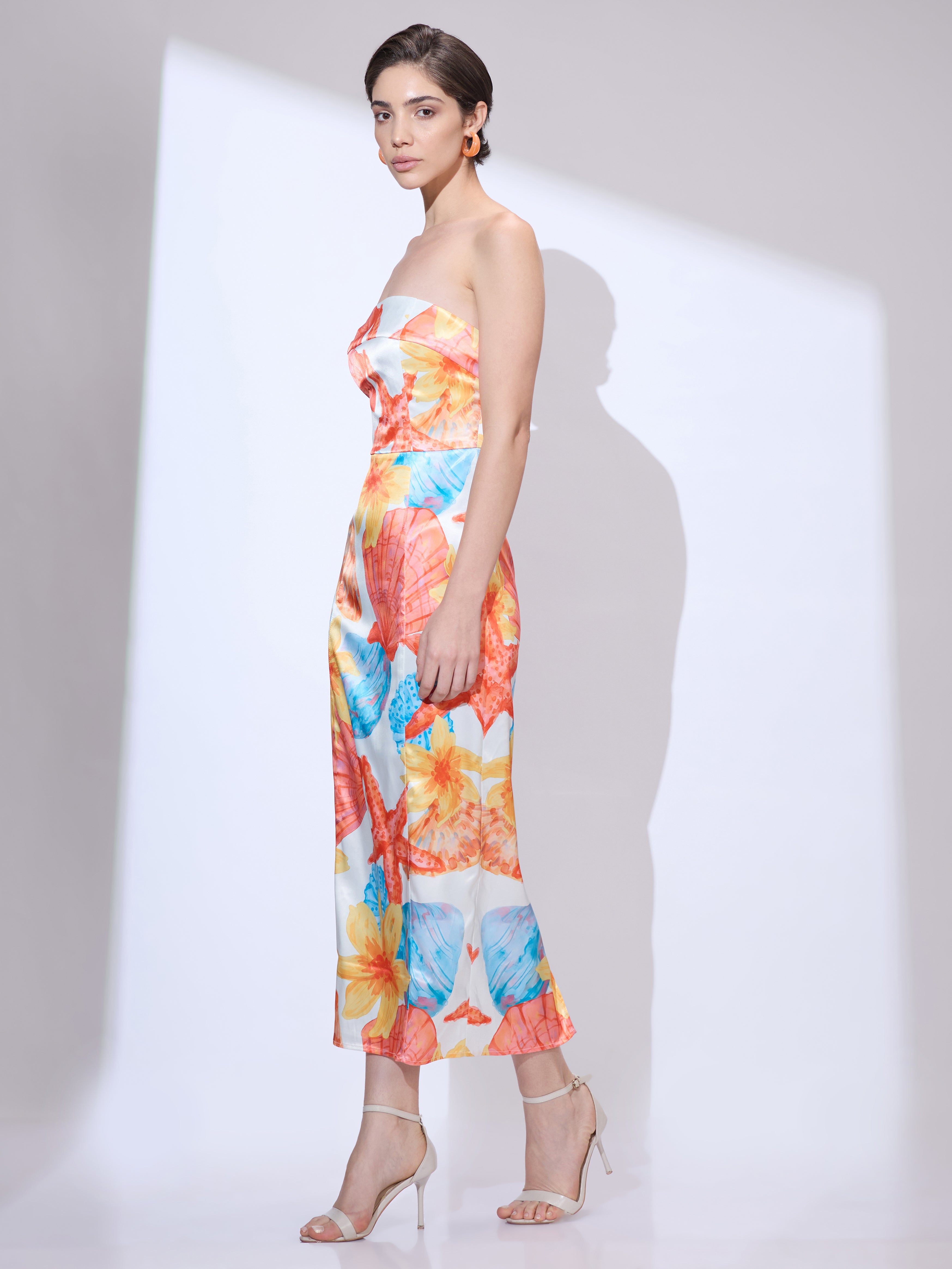 Image of Standing slightly to the side and facing forward in a colorful strapless midi dress from the resort wear collection featuring a seashell print