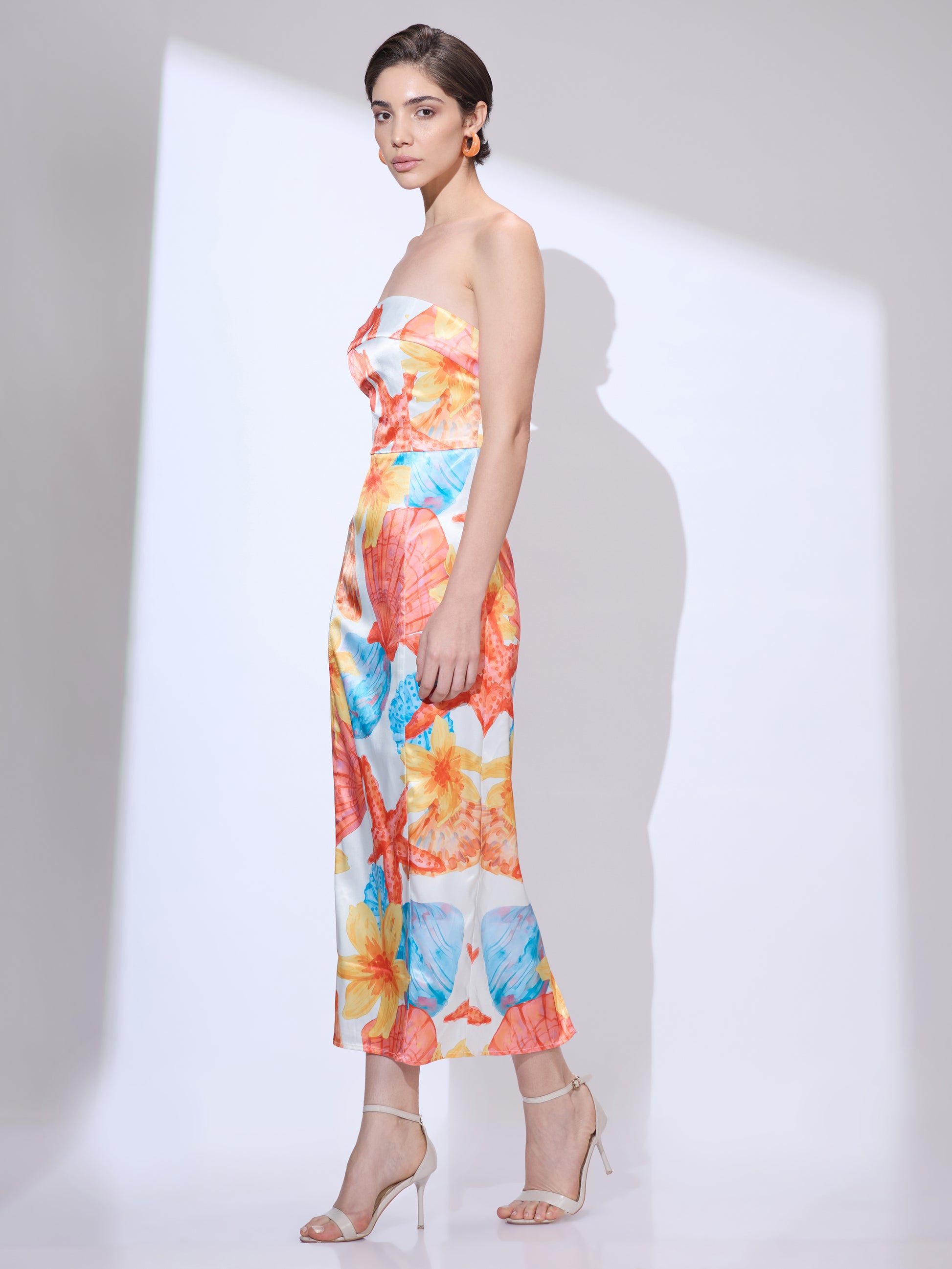 Standing slightly to the side and facing forward in a colorful strapless midi dress from the resort wear collection featuring a seashell print